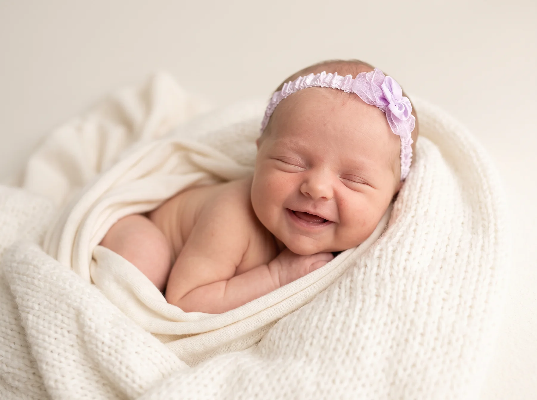 A smiling newborn wrapped in cream with a delicate floral headband
