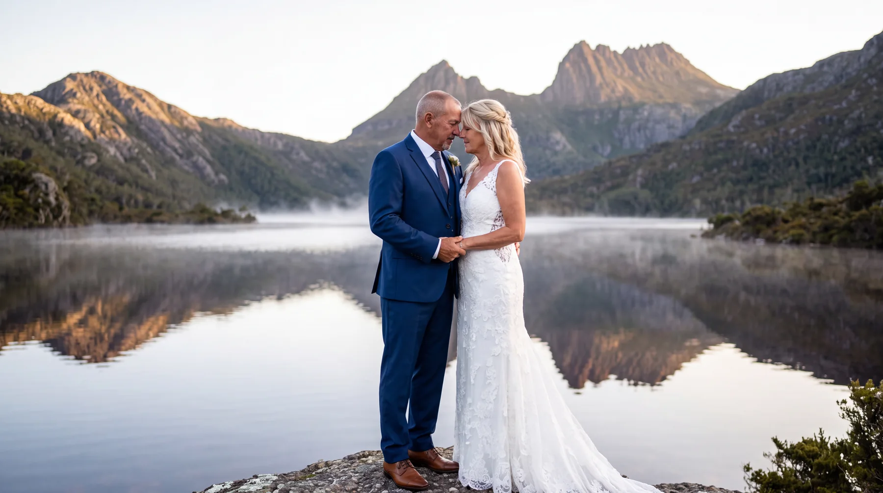 A bride and groom standing beside a reflective mountain lake